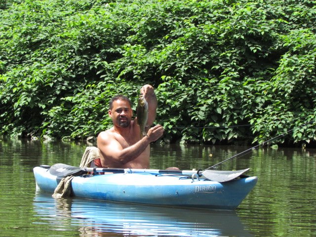 An angler holding a bass, river and trees behind him.
