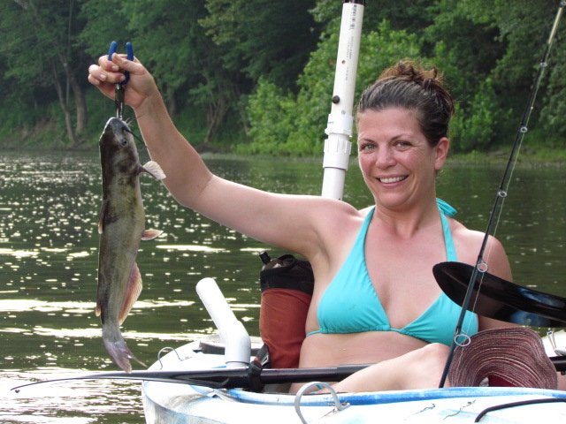 A woman in a kayak smiling and holding up a catfish.